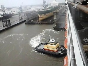 In this image taken from the Carnival Triumph (starboard side, looking aft), the towing vessel Noon Wednesday is seen taking position on the hull. The dry-dock facility and the still-attached work barges are also visible in this image.